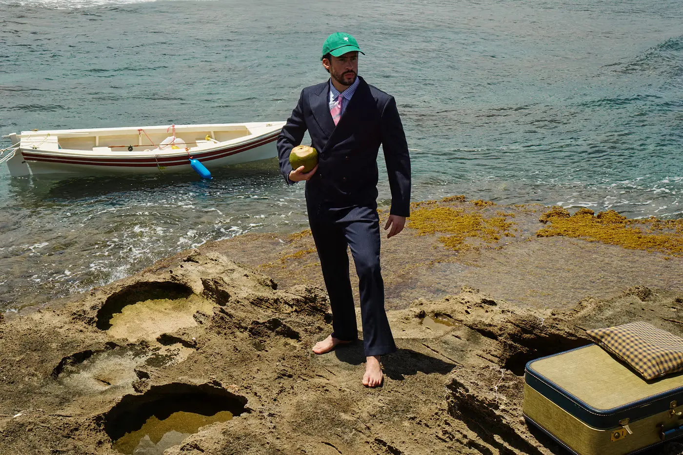 Bad Bunny porte un costume bleu foncé avec une casquette verte et tient une noix de coco, pieds nus sur des rochers dans un lieu côtier de Porto Rico.