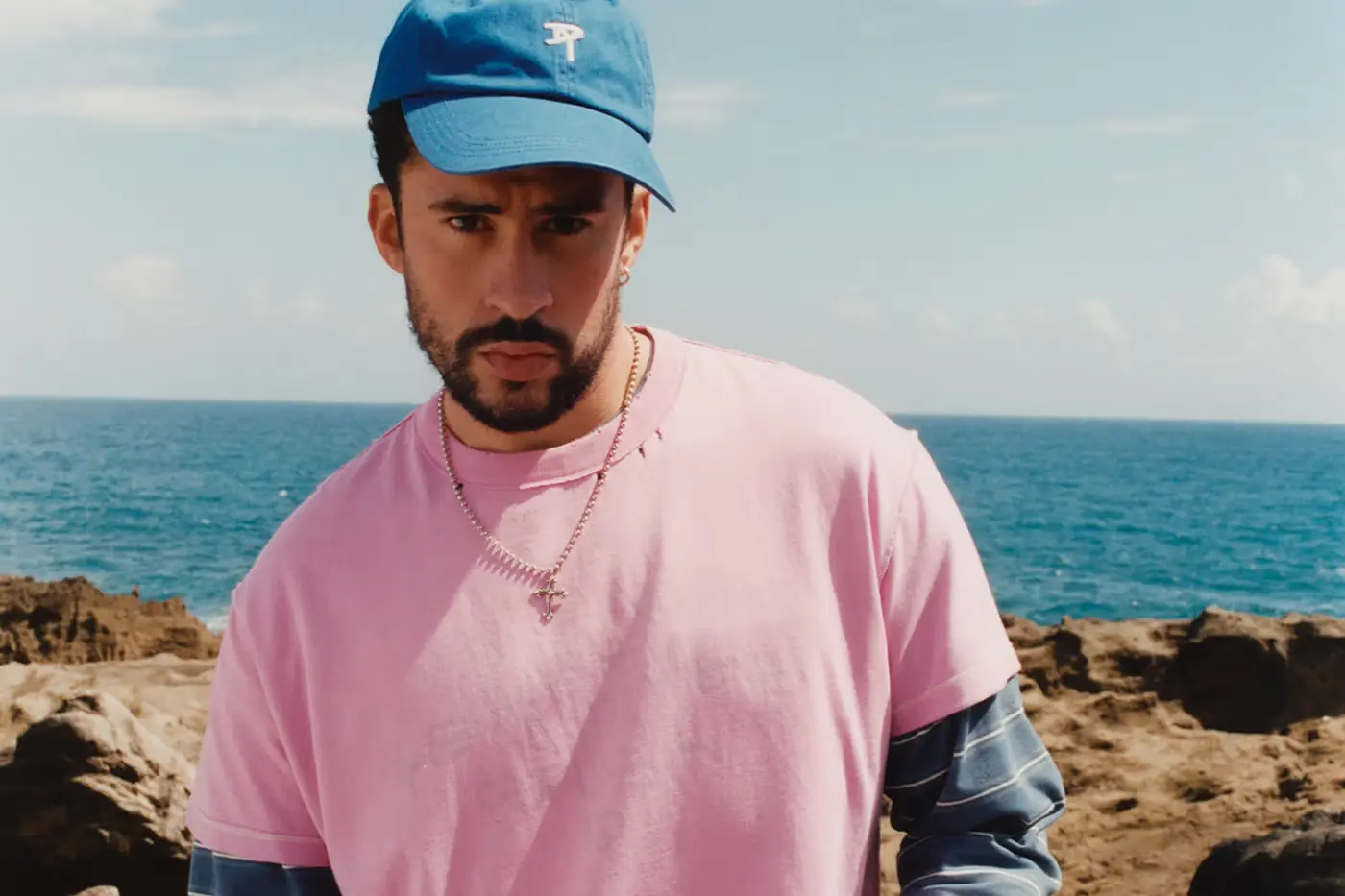 Bad Bunny porte un t-shirt oversize rose avec une casquette bleue et une chaîne en or devant un décor marin à Porto Rico.