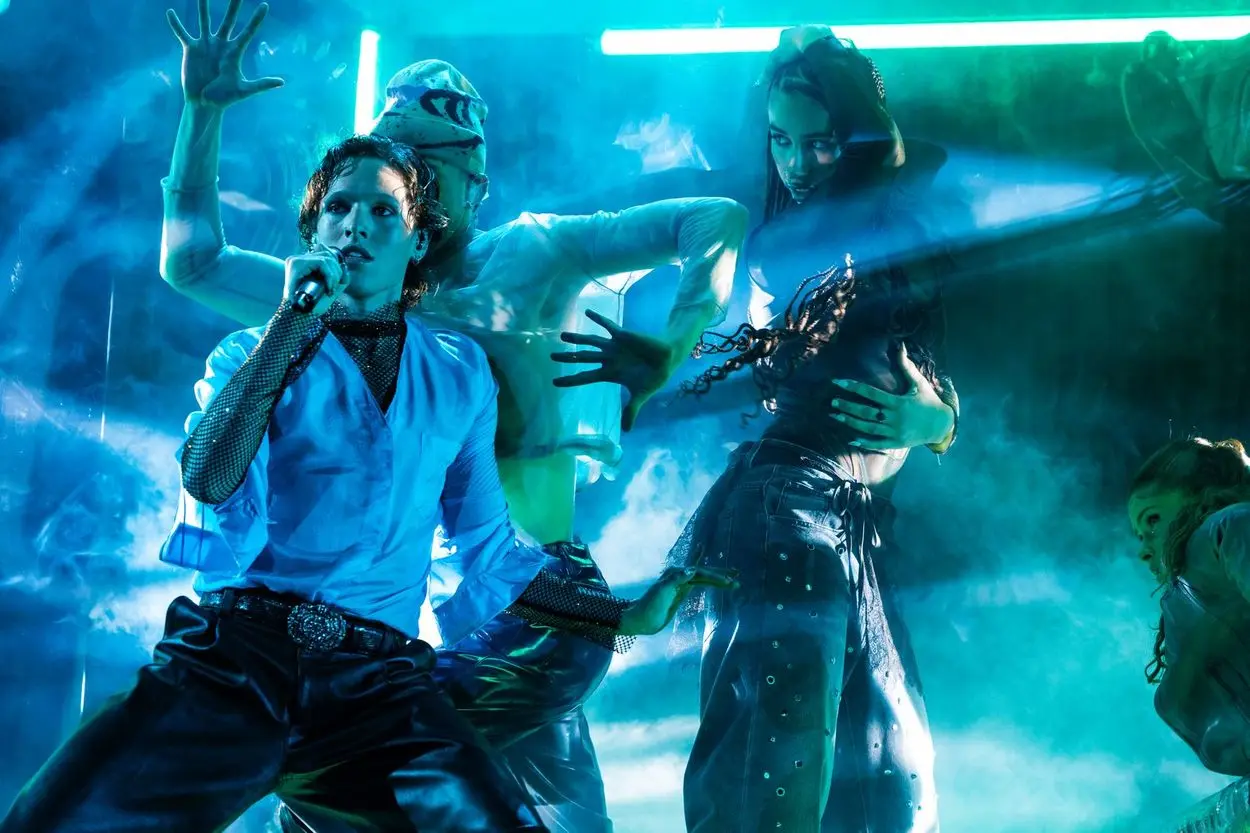 Søren Torpegaard Lund interprète «Før vi går hjem» pour le Danemark à l'ESC 2026 à Vienne: le chanteur en chemise blanche avec un détail en résille, entouré de danseuses et de danseurs dans une lumière de scène bleu-vert.