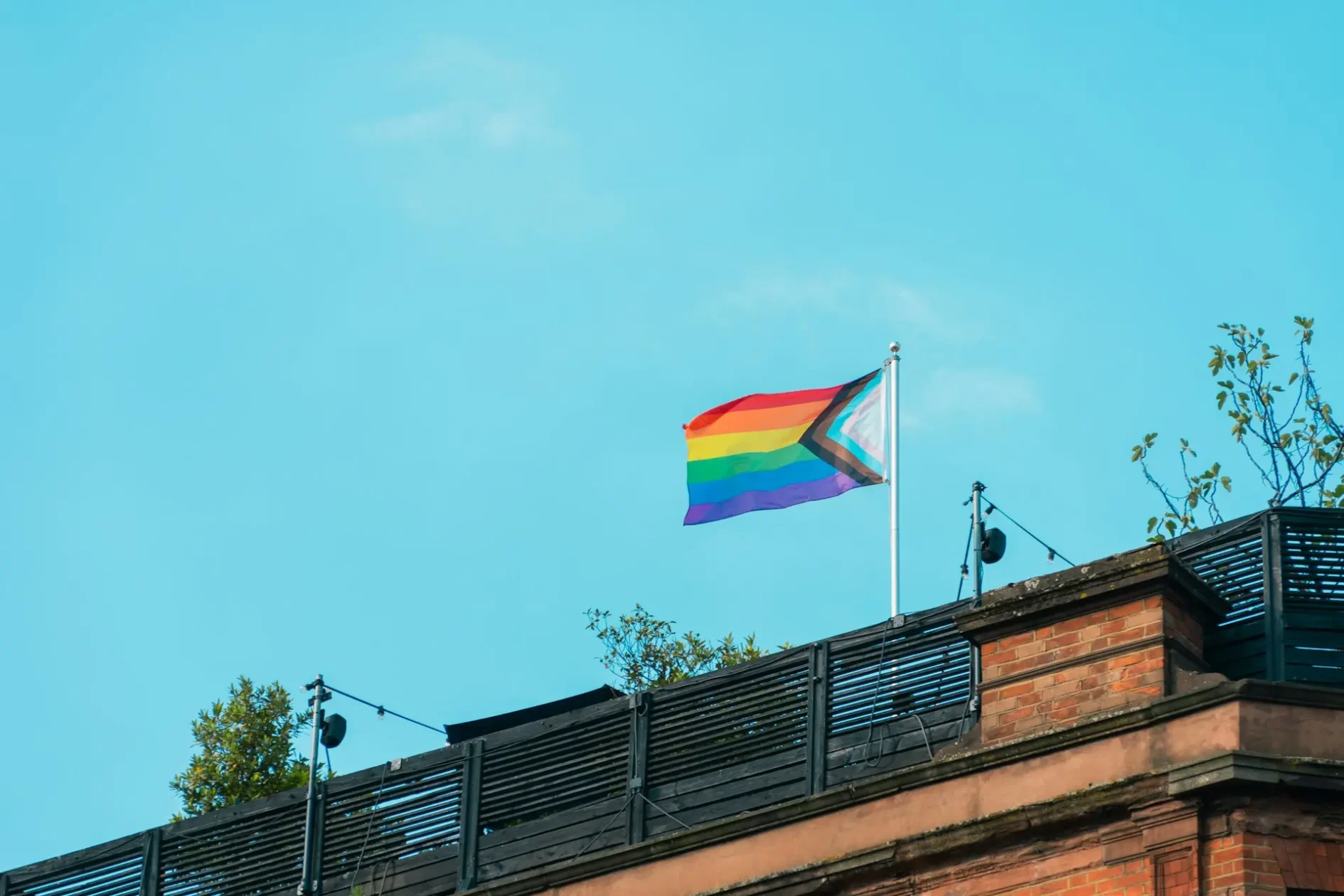 Progress Pride Flag weht auf dem Dach eines Backsteingebäudes vor blauem Himmel.