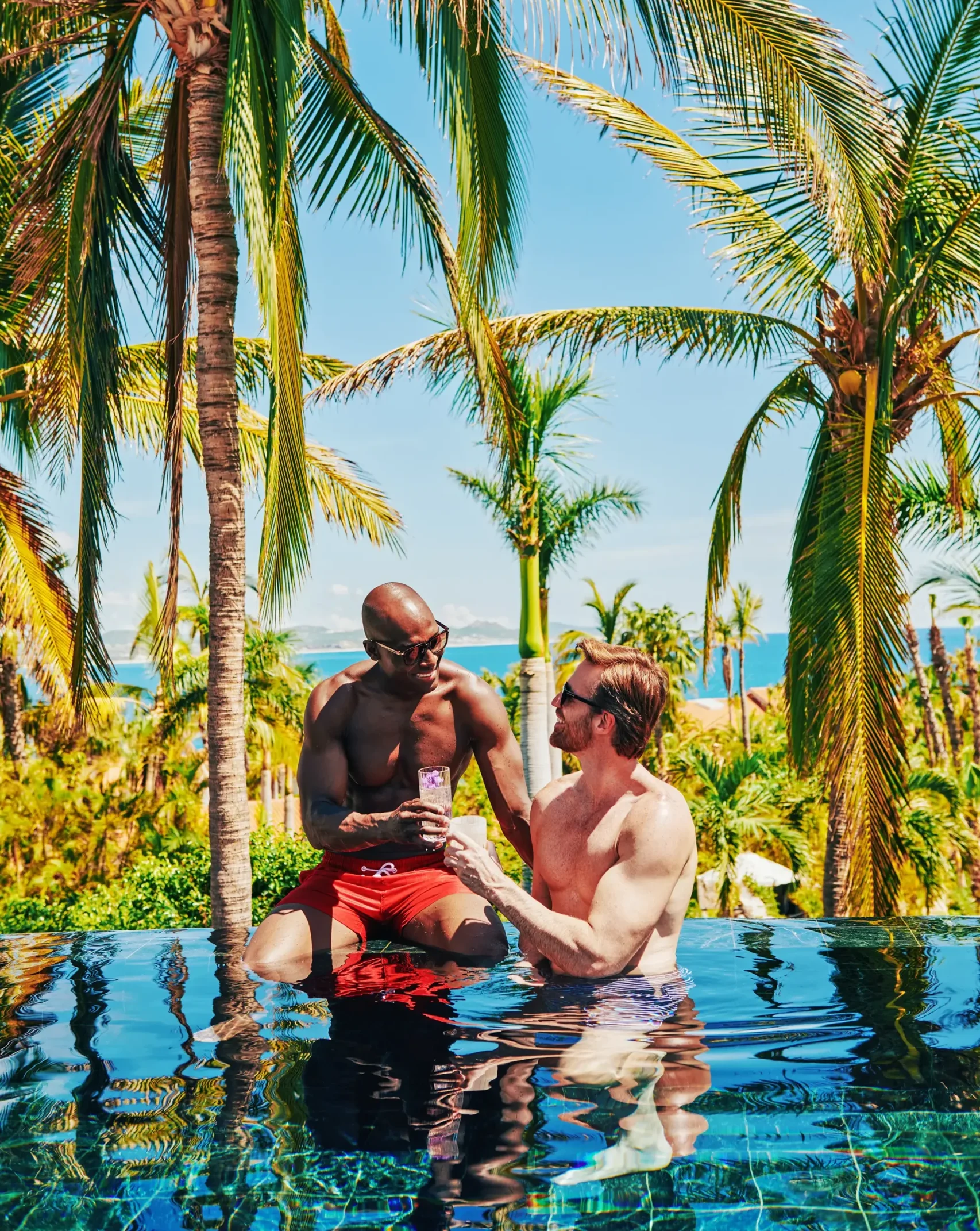 Deux hommes savourent un verre au bord de la piscine, avec des palmiers en arrière-plan.