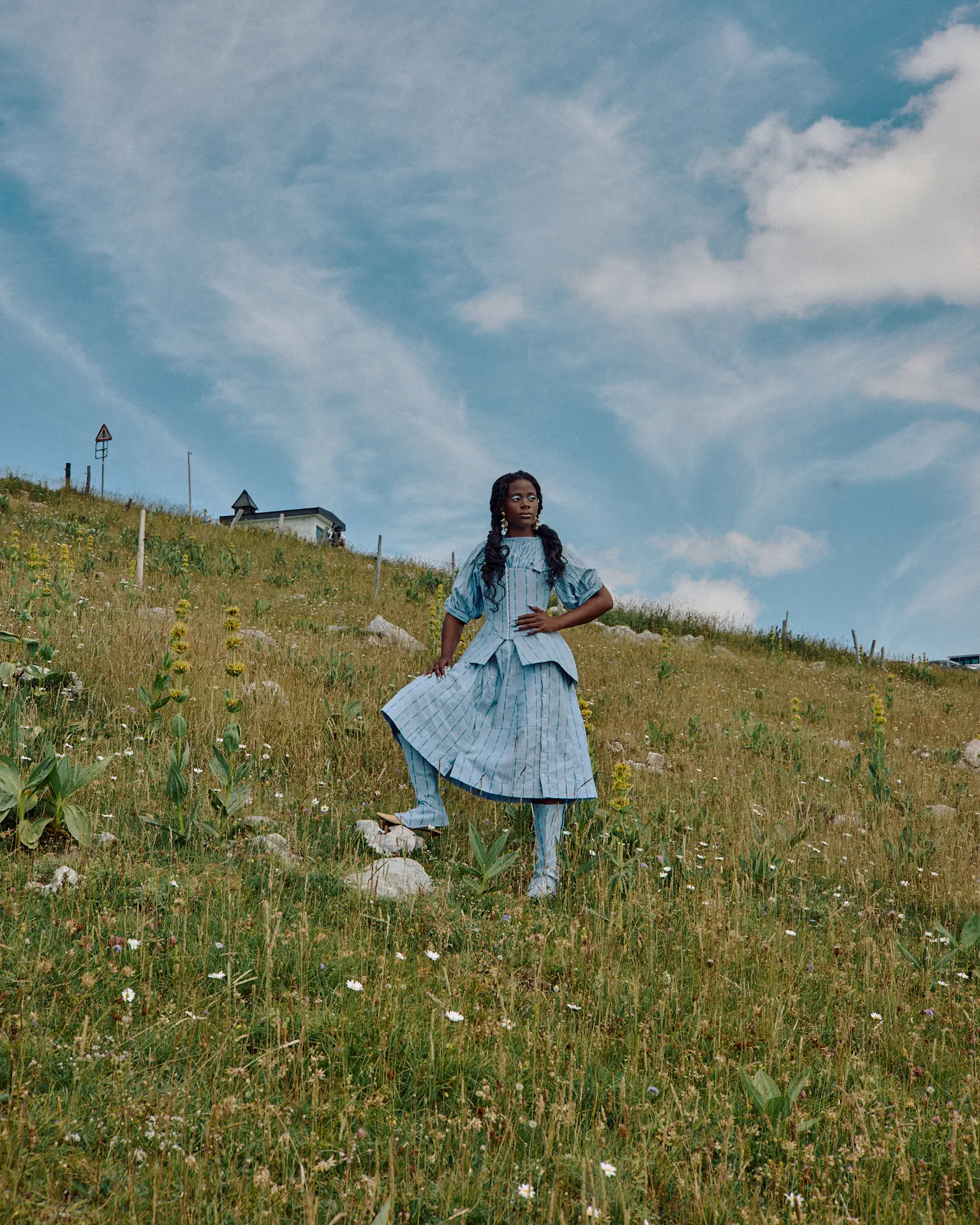 Eine Frau in einem blauen Kleid steht auf einem grasbewachsenen Hügel, wobei ein Fuß auf einem Felsen steht.