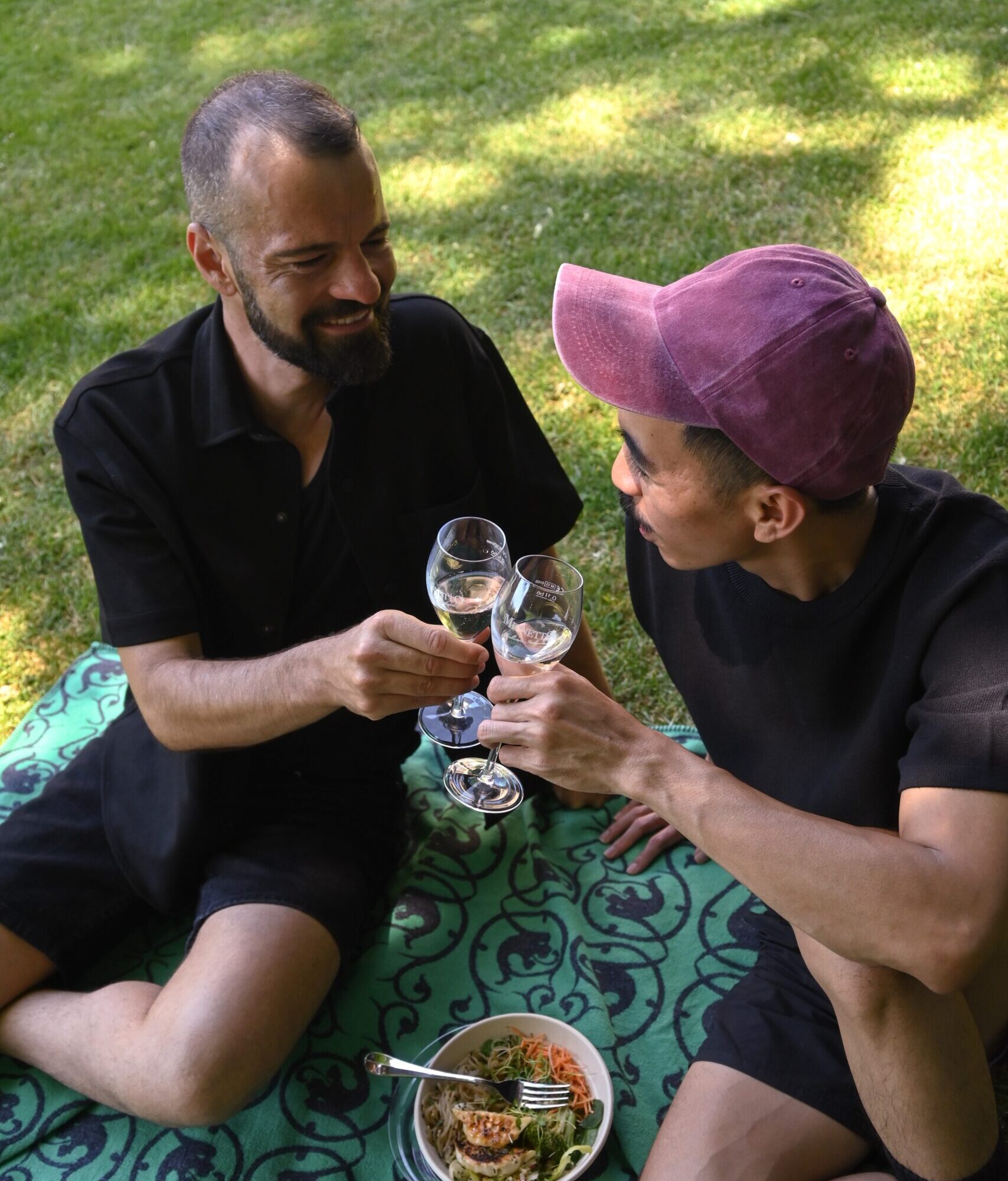 Zwei Männer stossen mit Champagner an, sitzen auf einer Picknickdecke, neben ihnen ein Korb voller Speisen.
