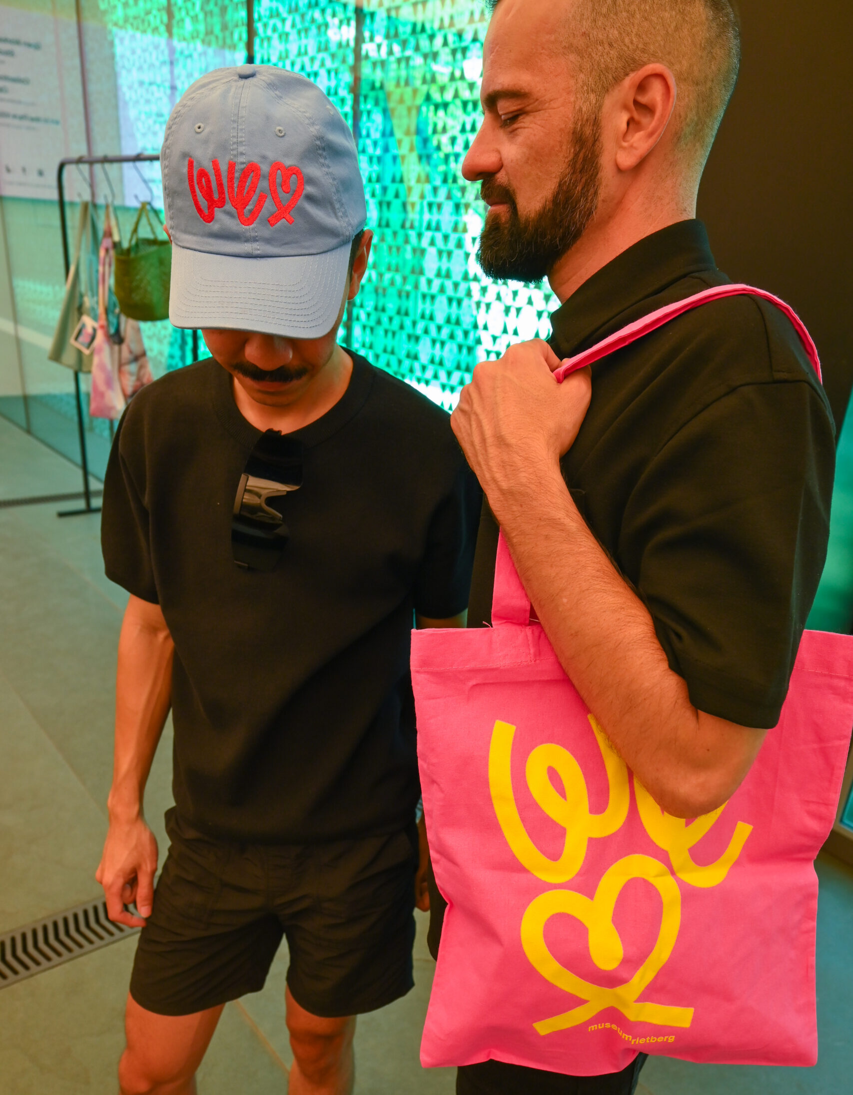 Zwei Männer tragen Merchandise-Artikel von WE LOVE. Der eine trägt eine pinkfarbene Stofftasche mit der Aufschrift „WE LOVE“, der andere eine blaue Kappe mit demselben Aufdruck.
