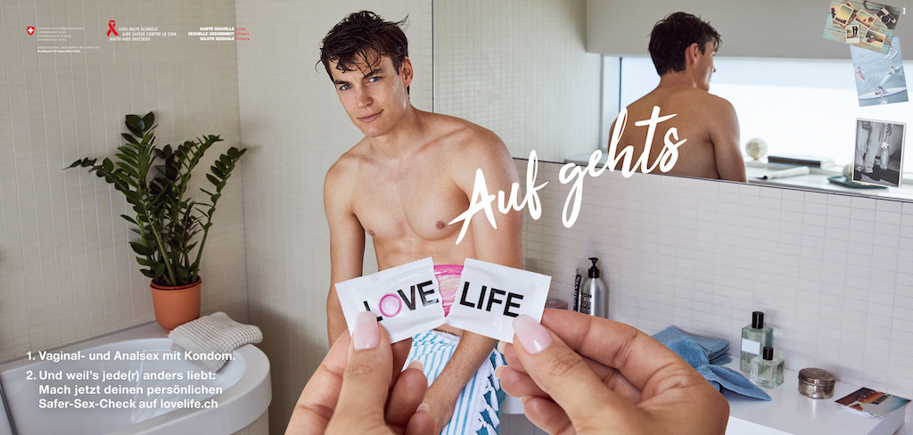 L'image montre un homme torse nu, appuyé contre un mur de salle de bain. Une paire de mains, plus proche de la caméra, ouvre un préservatif portant l'inscription «Love Life». Au-dessus du préservatif, en lettres grasses: «C'est parti». En bas à gauche suit le texte: «1. Sexe vaginal et anal avec préservatif. 2. Et parce que chacun·e aime différemment: fais dès maintenant ton propre Safer-Sex-Check sur lovelife.ch»