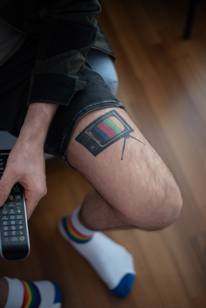 L'image montre Alexander avec une télécommande à la main et le tatouage d'une télévision vintage colorée sur sa cuisse.