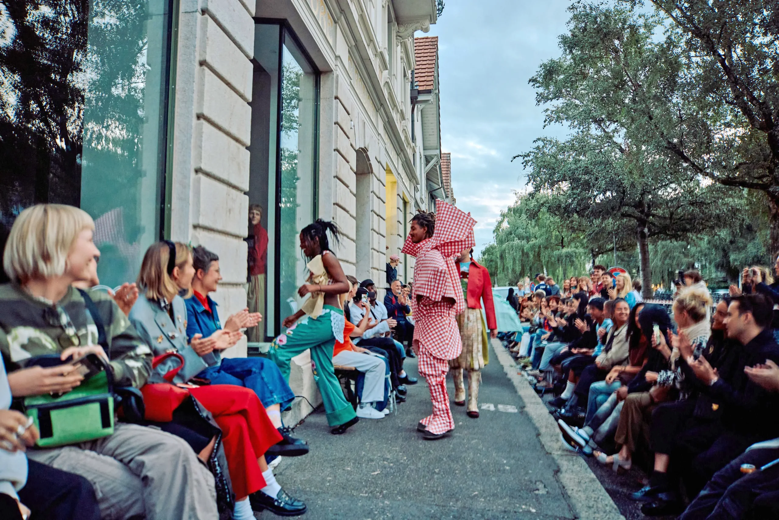 Le podium se trouve sur le trottoir d'une rue. À gauche, on voit les façades des bâtiments, à droite des arbres. Des spectateurs se sont rassemblés des deux côtés, et des mannequins défilent.
