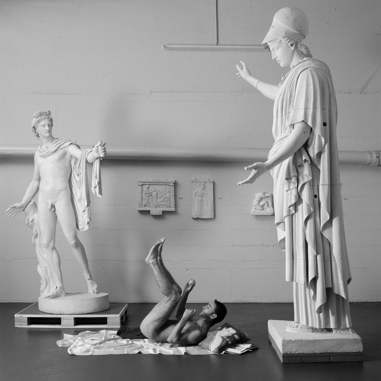 Un homme est allongé sur le dos et soulève les jambes, les bras et la tête du sol. Il est entouré de deux grandes statues. Il est allongé sur un drap et a un coussin sous la tête.