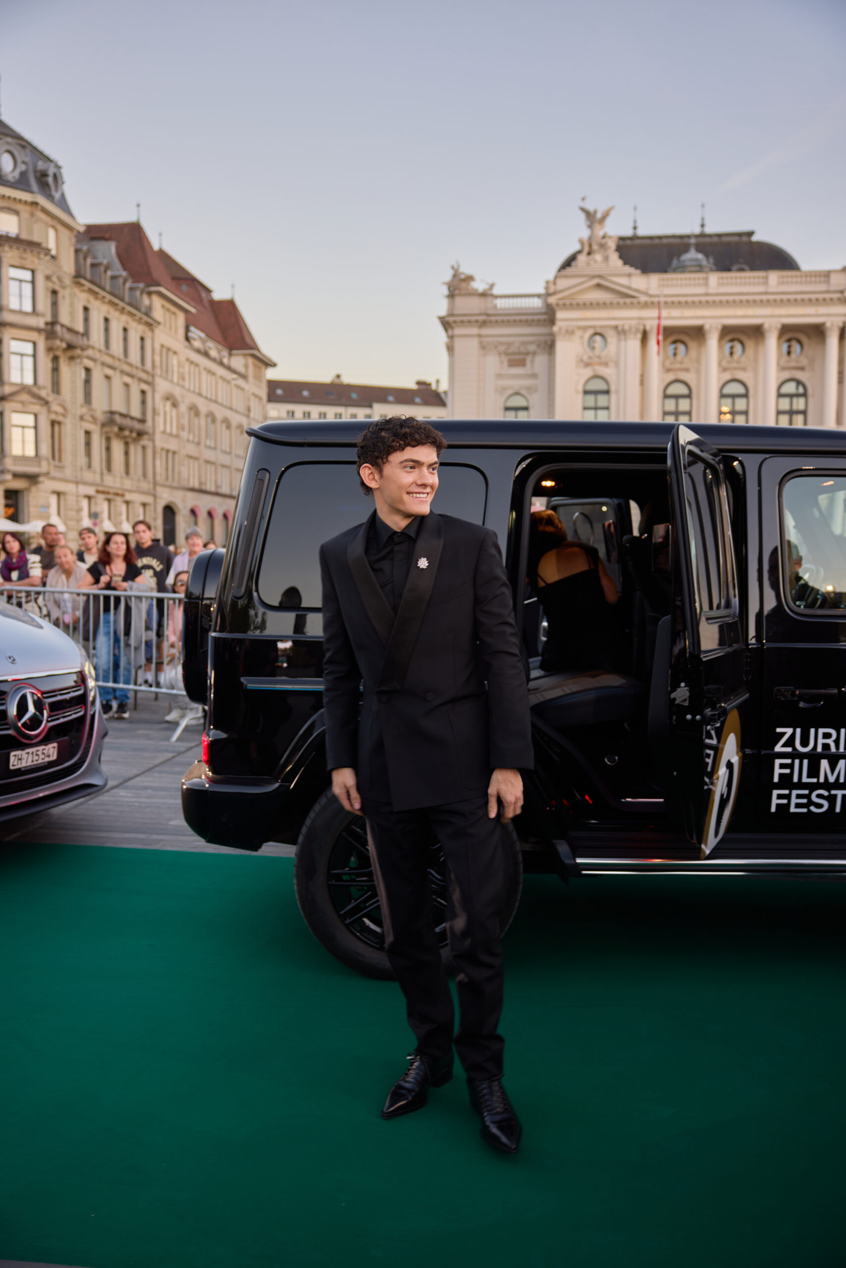 Cette image montre un homme debout à côté d'un véhicule noir dont la porte est ouverte, affichant le logo et le texte "ZURI FILM FEST" sur le côté. L'homme porte un costume noir et un nœud papillon, debout sur un green carpet. Derrière lui, une foule de personnes est rassemblée derrière une barrière, et une voiture Mercedes-Benz est partiellement visible sur le côté gauche. Dans le…