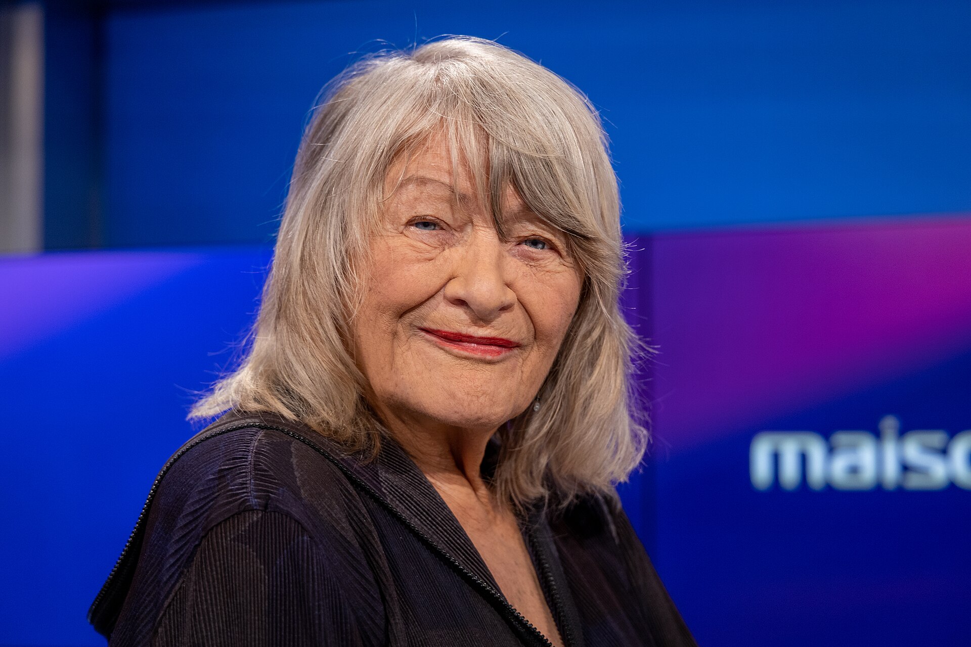 Alice Schwarzer mit grauem Haar lächelt in einem TV-Studio mit blauem Hintergrund