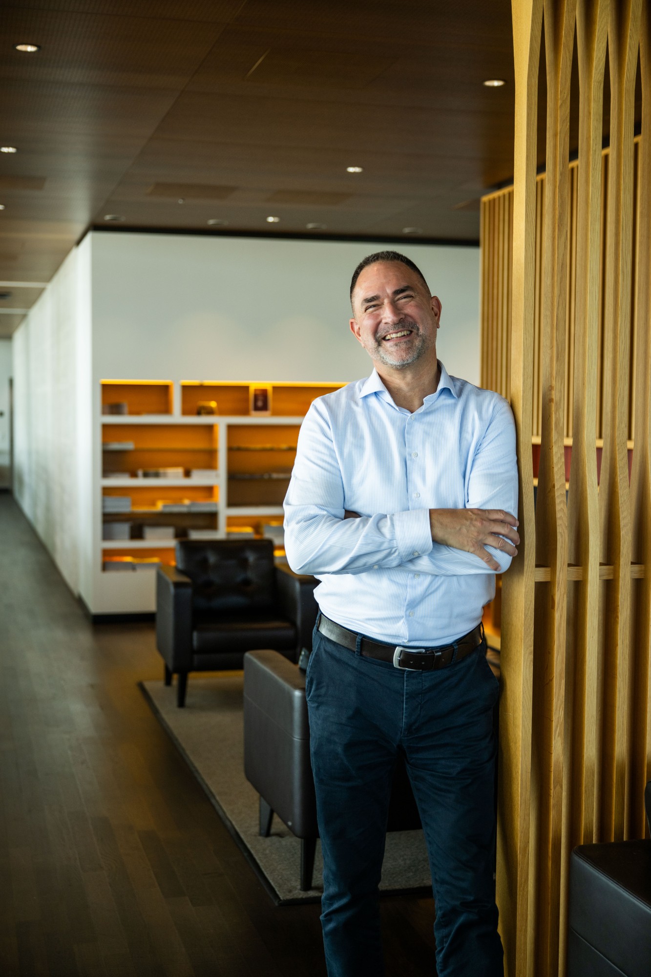 Roger Geu lehnt lachend mit verschränkten Armen an einer Holzlamellenwand in der SWISS First Lounge