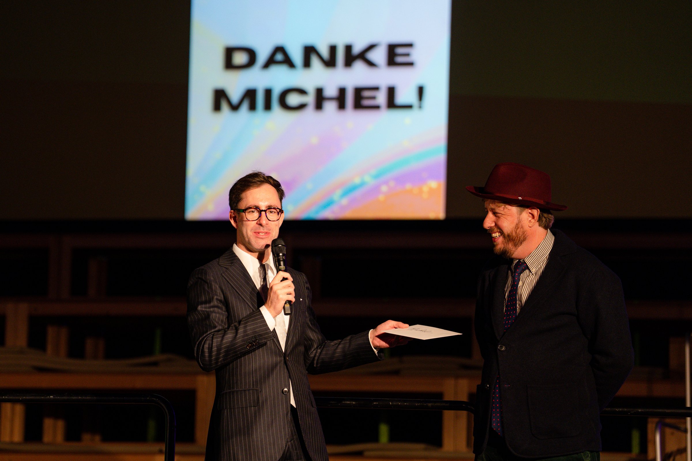 Zwei Männer auf der Bühne vor der Leinwand «Danke Michel!», einer überreicht einen Umschlag