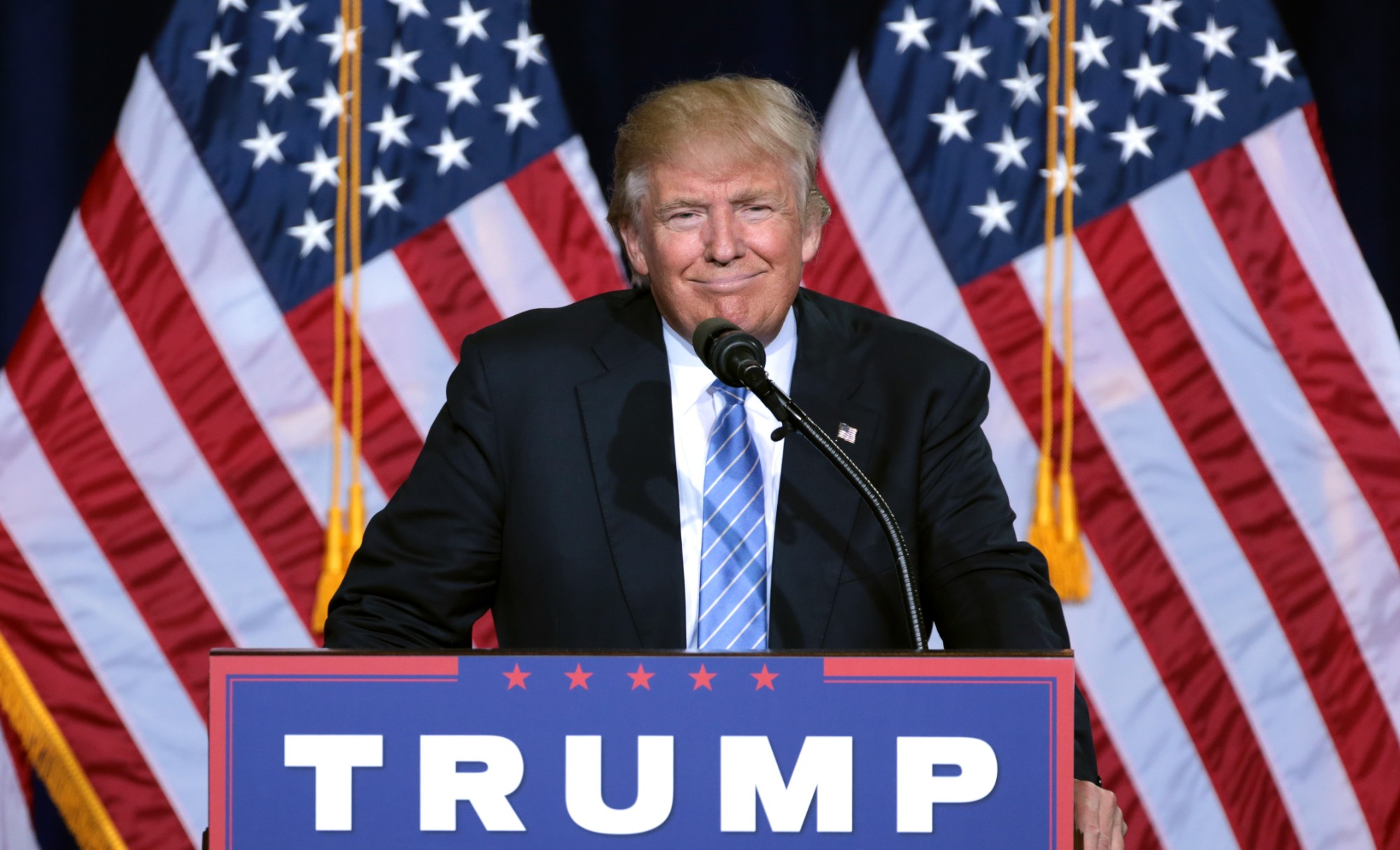 Donald Trump lächelt hinter einem Rednerpult mit der Aufschrift «Trump», hinter ihm zwei US-Flaggen