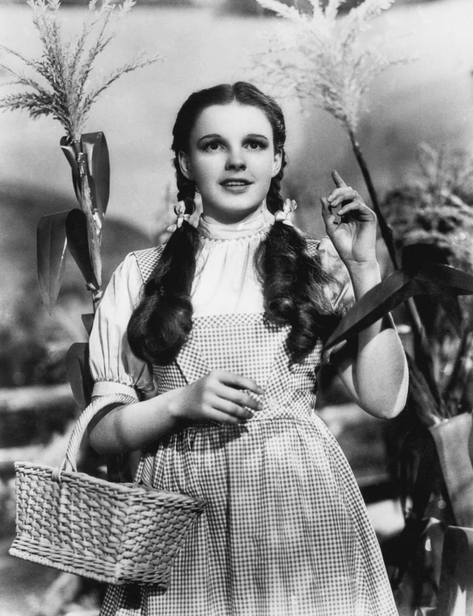 Schwarz-Weiss-Foto: Judy Garland mit Zöpfen, kariertem Kleid und Korb als Dorothy